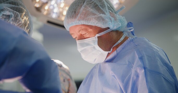 Close up of mature male surgeon in surgical suit operating patient with assistants in surgery room. Professional medics cauterize wound. Modern equipment in operating block. Medical staff at work.