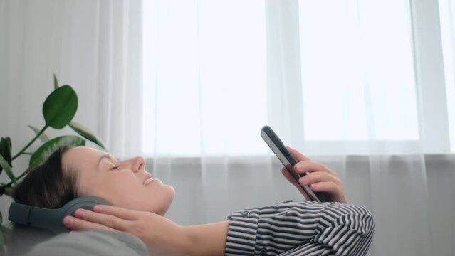 Peaceful happy young caucasian female wearing headphones, listening affirmations, visualizing future alone on comfortable couch. Charming tranquil woman listening slow pacifying music from mobile app