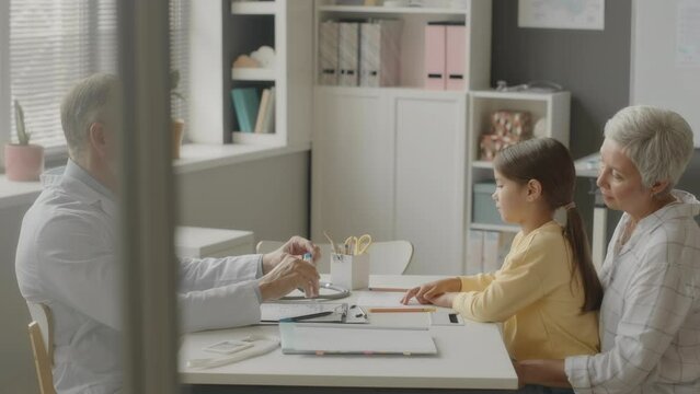 Side view medium slowmo of adult Caucasian male pediatrician wearing white lab coat giving thermometer to little Asian girl sitting on her grandmas laps in doctors office