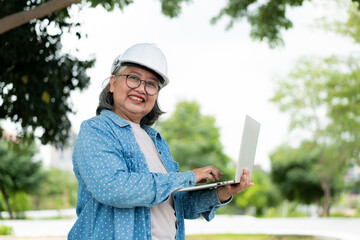 Happy elderly Asian professional engineer wears a helmet and checks the blueprint on a tablet on the construction site after the contractor and architect finish renovation the house and building