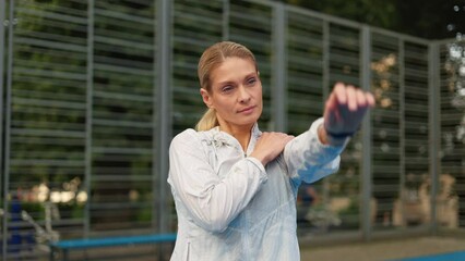 Middle-aged blonde trainer with pleasant appearance standing on court and performing arms exercises. Confident lady wearing white sport jacket, spinning hands in circle and later stretching them.