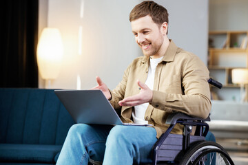 Handsome man with a disability sitting in wheelchair with laptop on knees and looking at camera. Caucasian male in casual wear having opportunities to remote work at home