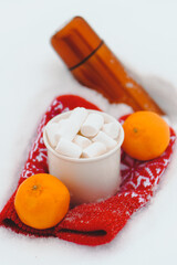 Christmas background with cup of marshmallow orange thermos and tangerines on red mittens on snow