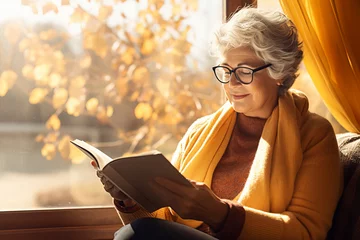 Foto op Plexiglas Beautiful senior lady reading a book by a window on sunny autumn day. Elderly woman enjoying nice fall weather. © MNStudio