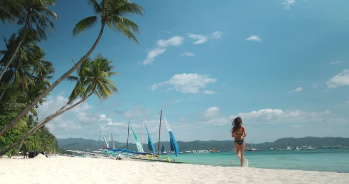 Long hair girl in bikini running white sand beach under palm trees on tropical Boracay island. People outdoor lifestyle travel, summer holiday vacation. Active sport, recreation. Slow motion back view