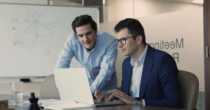 Multiethnic teammates using laptop met in office meeting room, mentor help with program to workmate, learn new software, share ideas, working on strategy, prepare on-line presentation. Apprenticeship