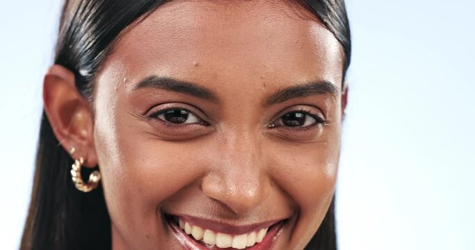 Face, funny and wink with a playful indian woman in studio on a blue background to flirt closeup. Portrait, emoji smile and a happy young person laughing at a meme or joke for freedom and comedy