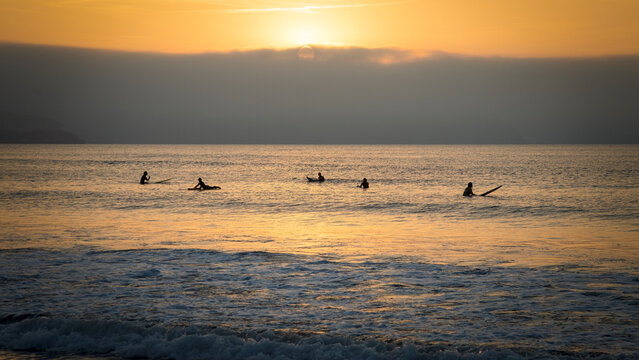 Atardecer dorado en una playa con la mar en calma y siluetas de surferos en el agua esperando olas que no vienen