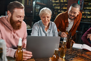 Fototapeta na wymiar happy multicultural colleagues looking at laptop while sitting with beer bottles in pub after work