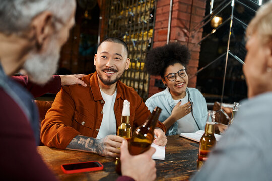 cheerful multiethnic business colleagues holding beer bottles and talking about new project in pub
