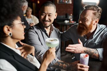 bearded tattooed man talking to amazed african american woman near smiling asian colleague in bar