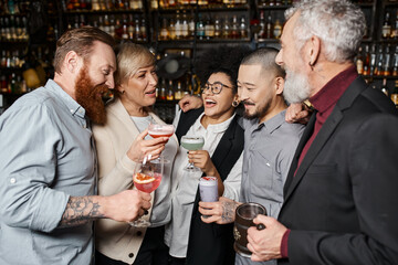 joyful multicultural colleagues holding glasses with drinks and talking in cocktail bar after work