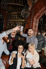 cheerful multicultural women looking at camera near bearded colleagues toasting in cocktail bar