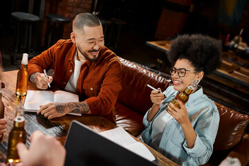 cheerful african american woman with beer bottle near asian colleague sitting with notebook in pub