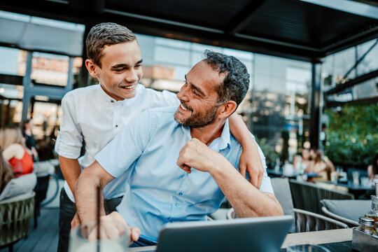 Handsome And Happy Father And His Teenager Son Sitting In A Restaurant And Talking.