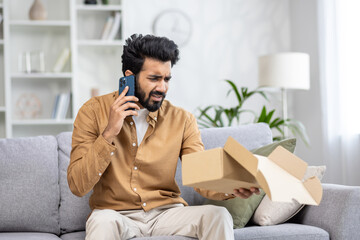 An angry young Indian man is sitting on the sofa at home, talking on the phone and looking disappointed at the open package he received.
