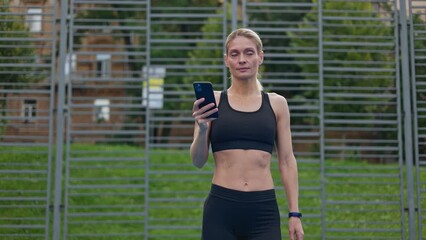 Confident young woman wearing black sportswear walking on court and looking at mobile phone screen. Slim blonde lady with gathered hair smiling and chatting with friend using online application.
