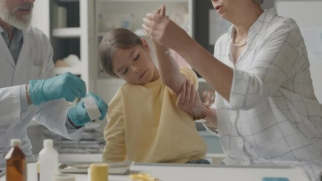 Medium shot of middle aged Caucasian male doctor wearing lab coat and medical gloves giving first aid to Asian little girl with insect bites on her arm