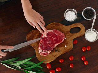 Woman with knife in hand cutting fresh steak meat for roasting kitchen with salt pepper other...