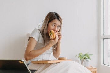 Asian Thai woman having breakfast on white bed while working, using tablet spending time at home alone on chilling day.