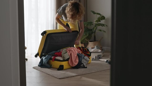 Mess before departure, woman shoves clothes into suitcase, dropping fussily in attempt to make it in time for departure. Female traveler chaotically fills suitcase, clothes sticking out of suitcase