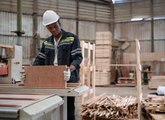 Young adult worker standing in warehouse examining hardwood material for wood furniture production. Male supervisor wears uniform and safety hardhat working in lumber pallet factory. Woodwork industry