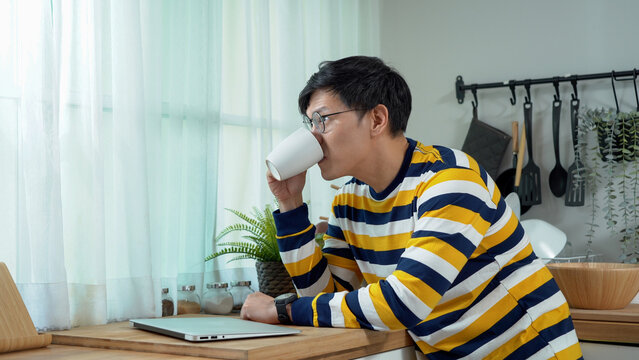 Asian Business Man Using Laptop And Drinking Coffee Looking Out Of Window In Kitchen Working At Home In Morning Light