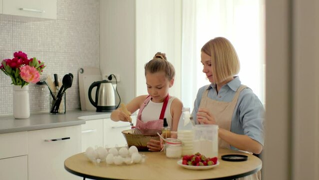 Mother and daughter are preparing pancake batter at the table in a modern kitchen. The girl holds a whisk, stirs eggs in a bowl, prepares dough with her mother.