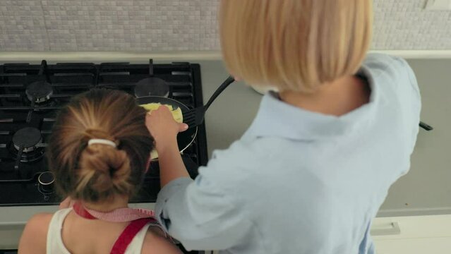 A young mother and her little daughter cook pancakes together for breakfast. Mother teaches daughter to cook.
