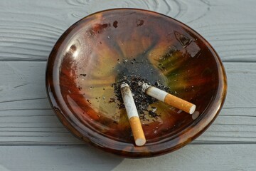 one beautiful shiny original glass brown round porcelain ashtray with two cigarette butts with a yellow filter and black ash stands on a wooden gray table during a meeting of friends