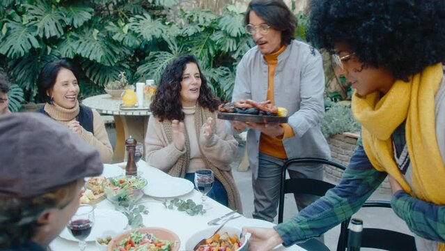 Young couple bringing dishes with grilled food to dinner table, joyous friends clapping hands and clinking drinks in a toast during BBQ party outdoors in the backyard