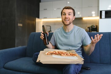 Rest after hard work. A man is sitting on the sofa at home, eating pizza and watching TV.