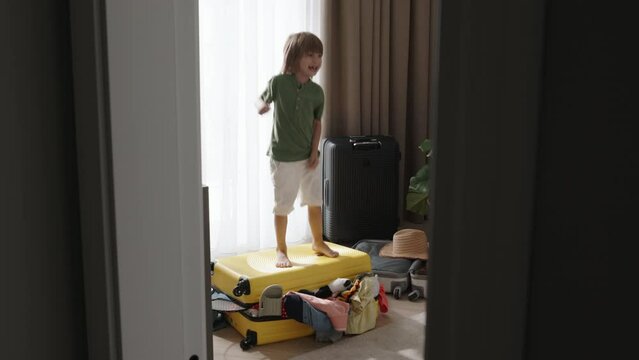 Boy Joyfully Jumps On Packed Suitcase. Energetic Happy Child Prankster Troublemaker Made Mess In Perfectly Packed Luggage Jumps On Suitcase. Messy Stacked Clothes In Luggage Sticking Out Of Suitcase