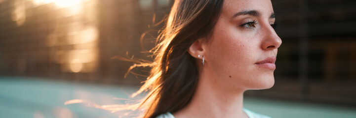 Close-up portrait of young woman with freckles and dark loose hair and long eyelashes wearing white top looking straight at the camera. Beautiful girl on modern city background