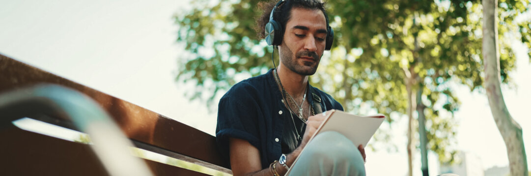 Fototapeta Young italian guy with ponytail and stubble sits with headphones on street bench listens to music and makes sketches with pen on piece of paper on cityscape background
