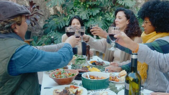 Cheerful mature woman saying drinking toast and raising wine glasses with friends while celebrating holiday at outdoor dinner party. Zoom out shot