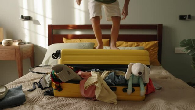 Child tourist jumps on packed suitcase. Delight from upcoming family trip to resort countries by plane. Tourist packed baggage with clothes, toys sticking out. Boy kid bouncing on yellow suitcase.