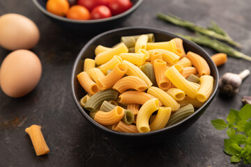 Rigatoni colored raw pasta with tomato, eggs, spices, herbs on black concrete. Side view, selective focus.