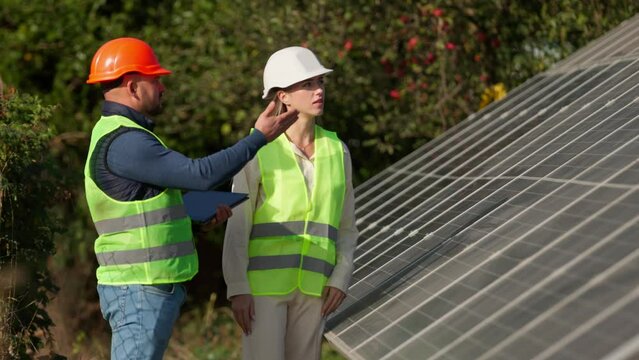 A man and a woman in hard hats and with a laptop are discussing work tasks against the backdrop of solar panels outside. Female environmental engineer talking to investor. Green electricity concept.