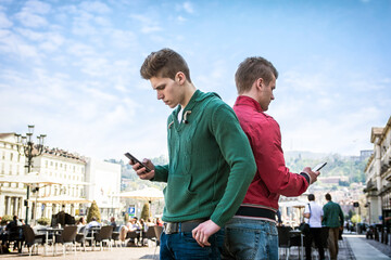 A couple of men standing next to each other, back to back, engrossed in their cell phones and ignoring each other's company - obrazy, fototapety, plakaty