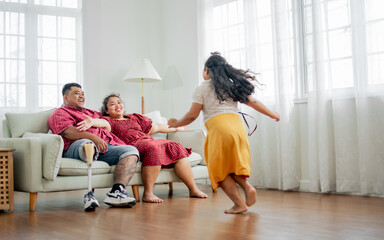 Selective focus Asian happy father with disable prosthetic leg and mother sitting on sofa at cozy home, raising to hug little daughter girl with love and warmth. Family, Interior, Healthcare Concept. - obrazy, fototapety, plakaty