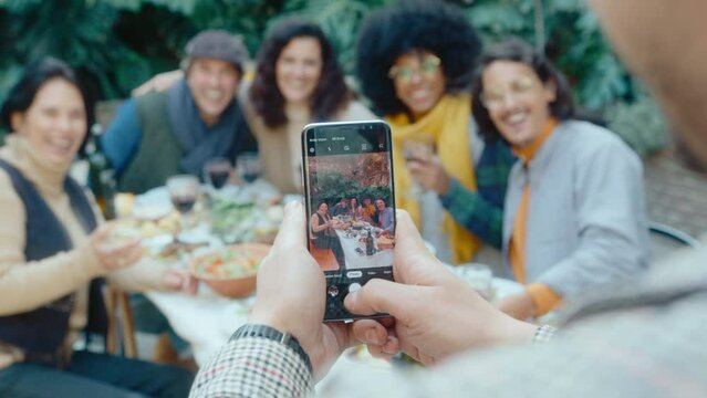 Man taking photos with phone of happy friends sitting around dinner table with food and drinks on outdoor party in backyard. Rack focus shot