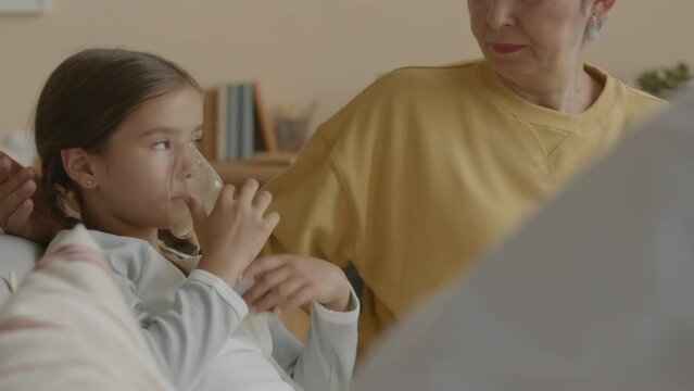 Waist up of Asian little girl breathing through inhaler while sitting on couch with caring mother speaking to doctor during home visit