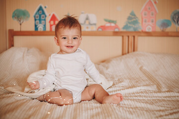 Fototapeta premium A little baby in a white bodysuit sitting on a bed and smiling