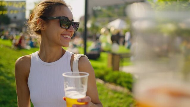 POV happy joyful young woman have fun at outdoors picnic on green meadow. woman holding plastic glass with beer and cheers with someone. party time outdoors. Happy hour, lunch break and youth concept