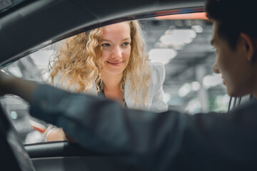 A beautiful female employee of a car rental shop gives documents to a handsome young man to sign, accepting the terms of the car rental agreement, sitting in the car.