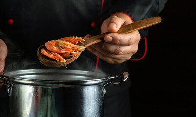 Professional chef prepares delicious shrimp in a pan in the kitchen. The concept of a delicious shrimps breakfast for a hotel. Spoon in the cook hand wis scampi - obrazy, fototapety, plakaty
