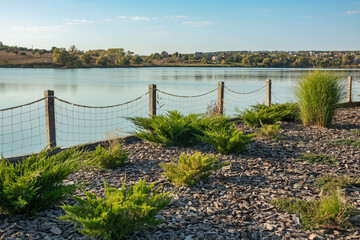 Landscape design with bushes on the shore of a lake with a fence made of ropes and wooden posts