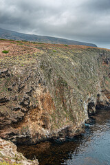 Santa Cruz Island, CA, USA - September 14, 2023: Covering rock fell partly off due to surf and storms revealing brown spots. Caves at water level. Gray cloudscape. Sparse green spots