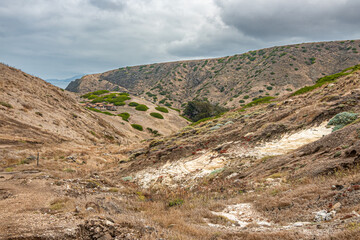 Santa Cruz Island, CA, USA - September 14, 2023: Rough hilly dry landscape with some green patches and one large white exposed hard stone site under storm cloudscape